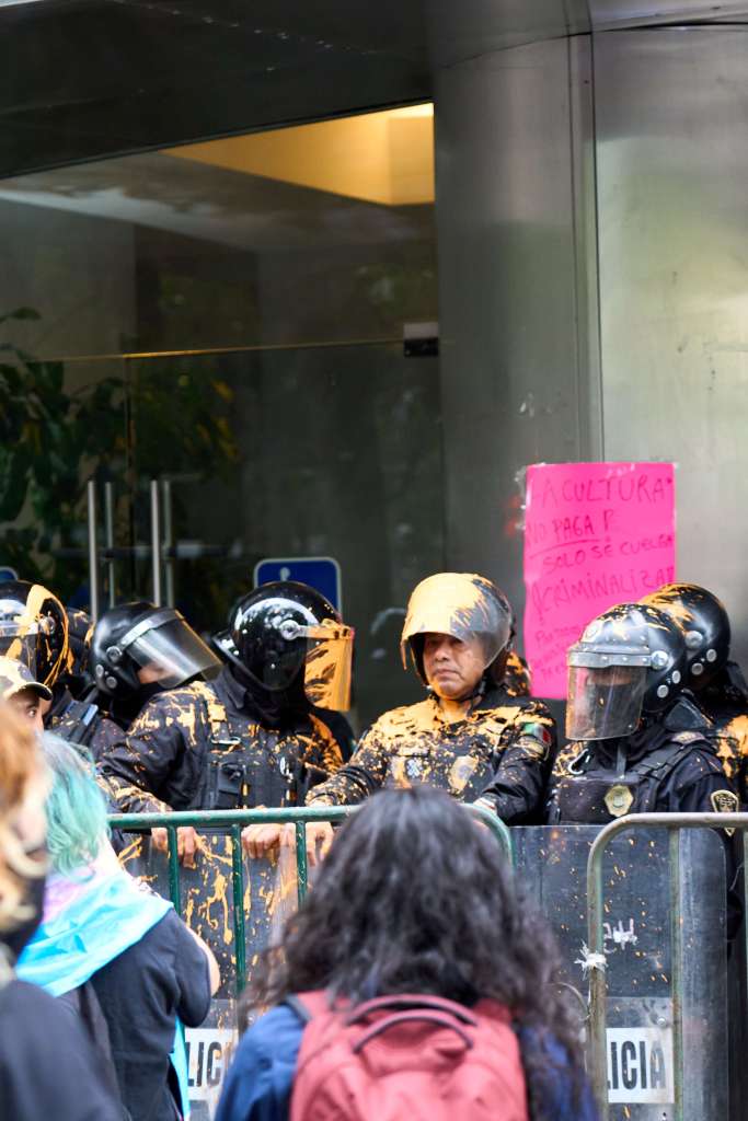 Police officers stand miserably behind a barricade, covered head to toe in yellow paint.