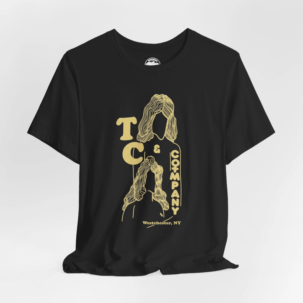 .A photo of a black t-shirt with the TC & Company logo on it, featuring yellow stensiled outlines of two silhouetted women, one overlaid above the other.