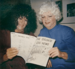 Two transfemmes in wigs reading an issue of The Toronto CDC News, giving major 90s lesbian vibes.